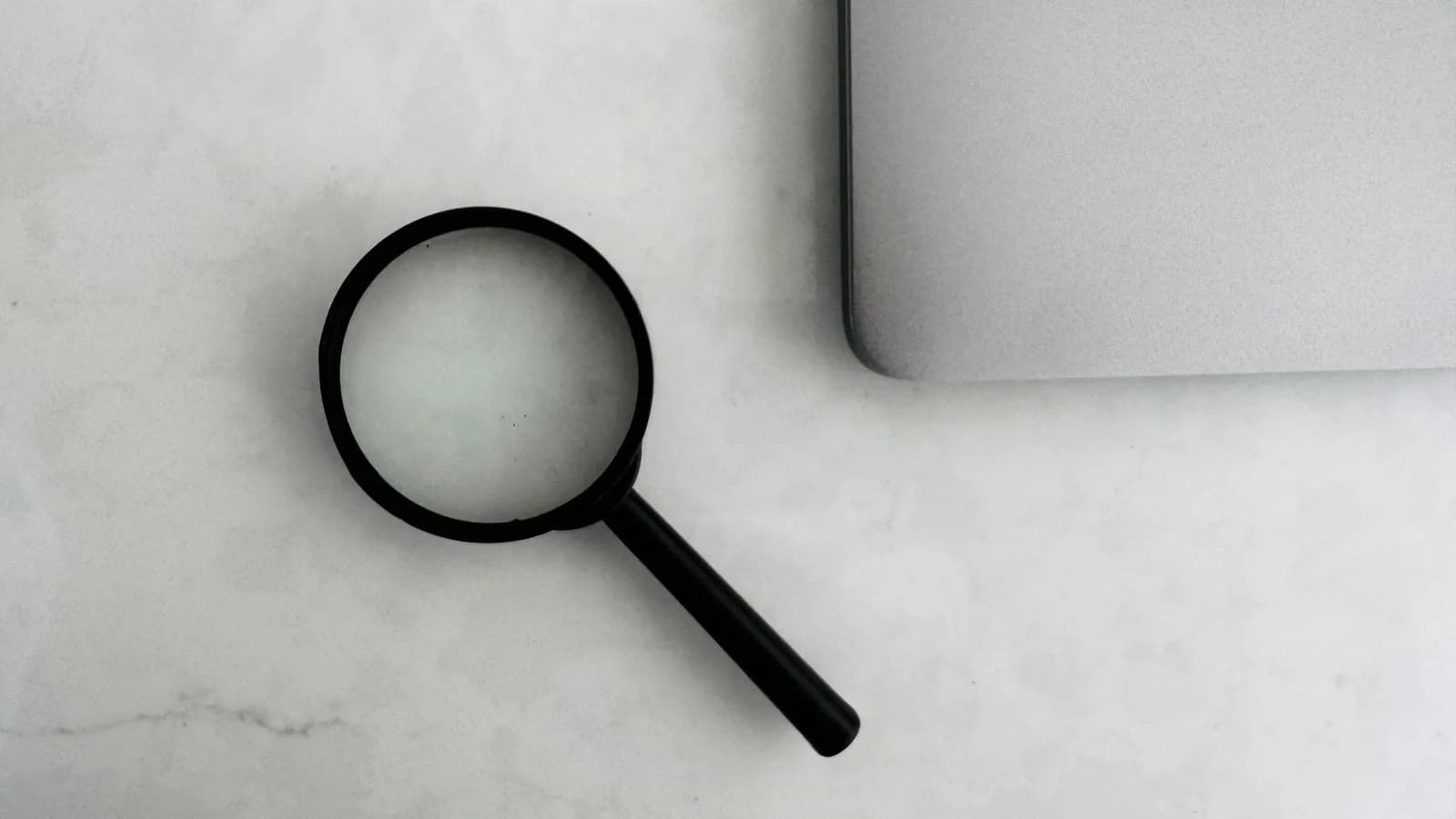 A magnifying glass beside a laptop on a light marble surface