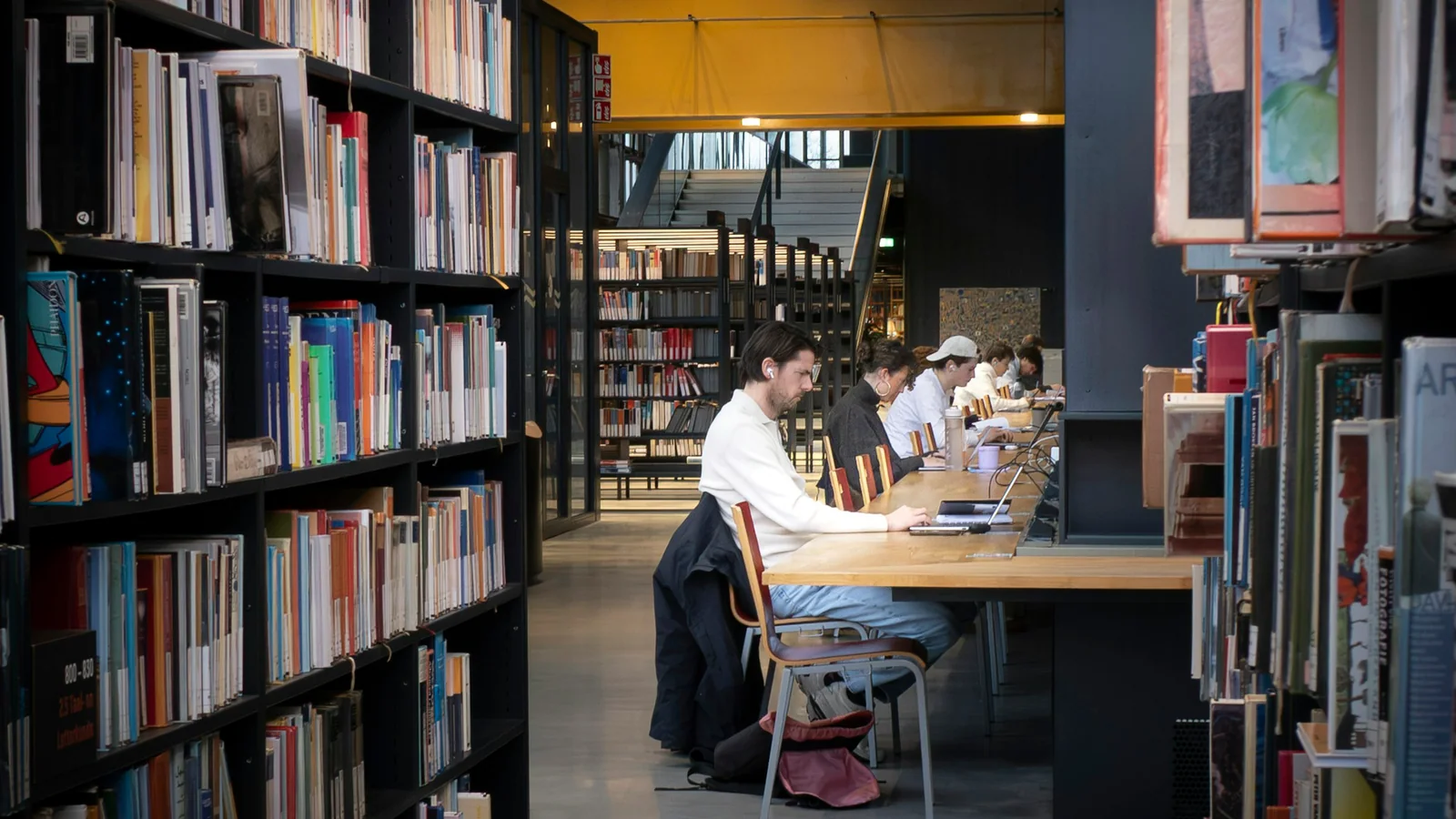Students studying at long wooden tables between library bookshelves