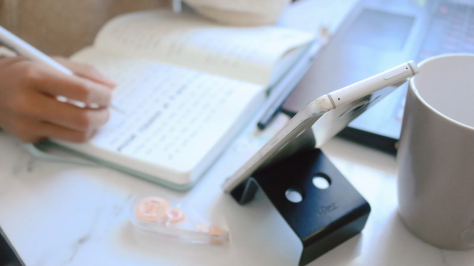 A hand writing in a notebook beside a tablet at a white desk
